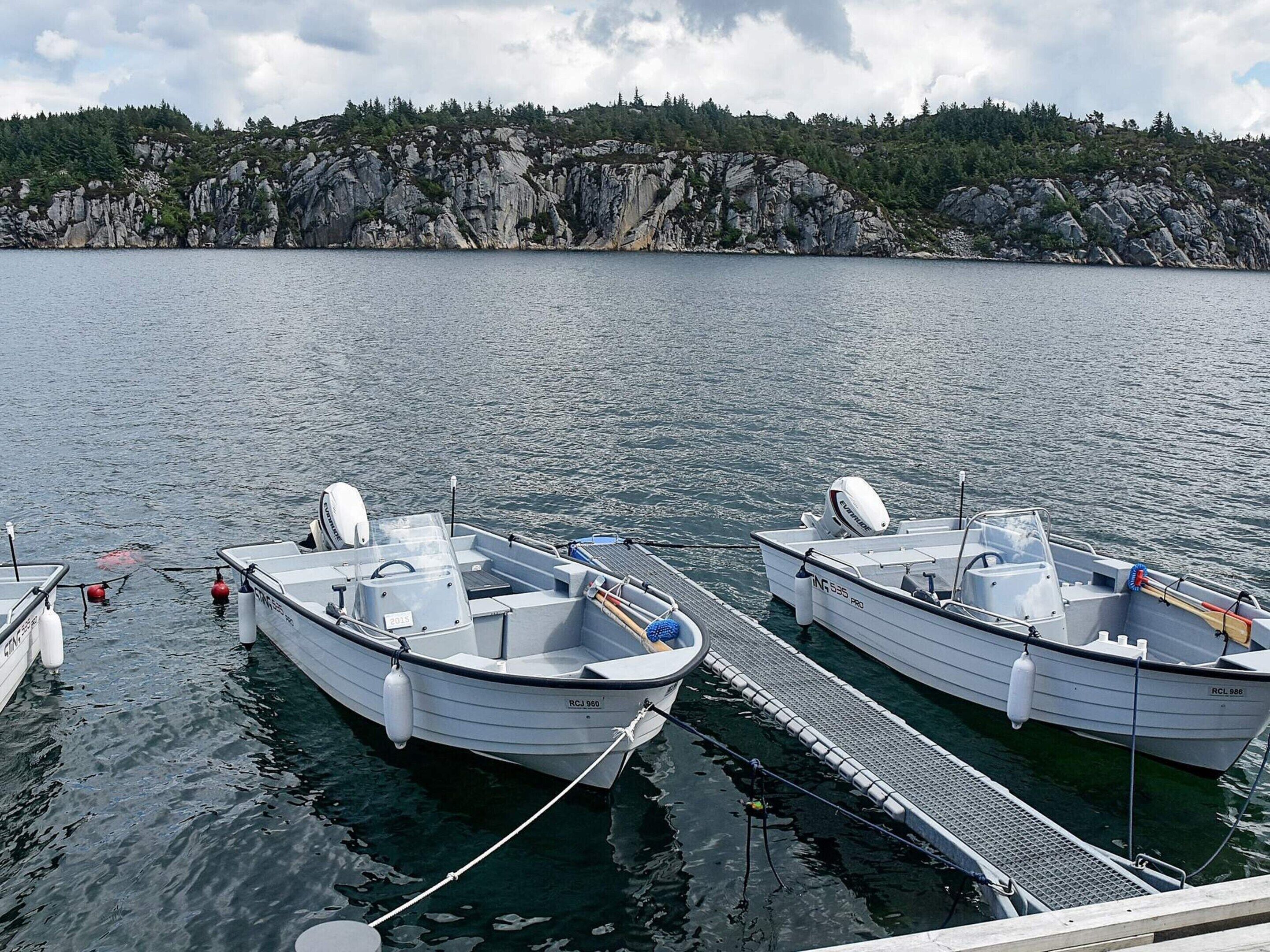 Dois pequenos barcos a motor atracados num cais com uma costa rochosa e colinas arborizadas ao fundo.