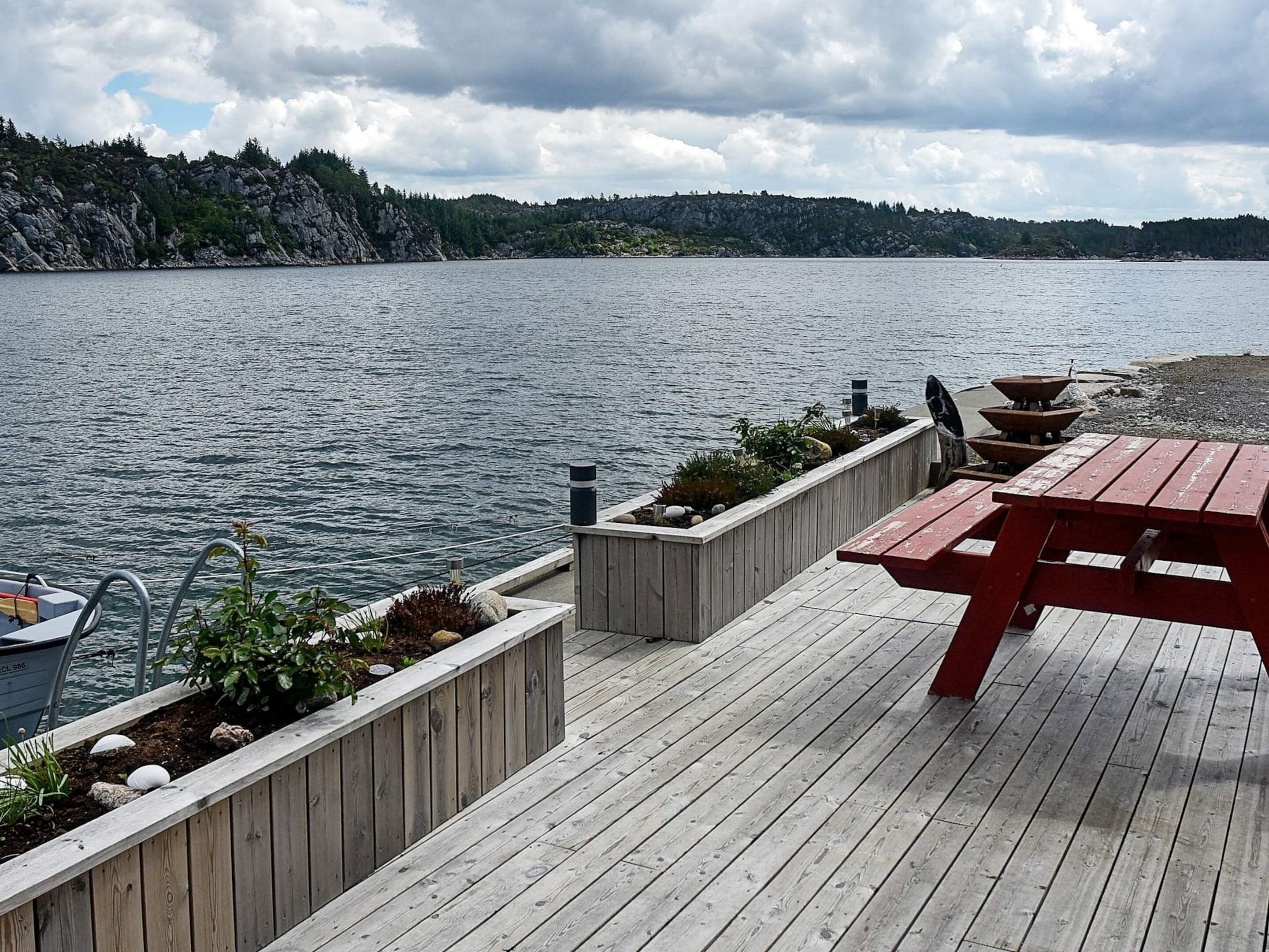 A wooden dock with a picnic table and planters, overlooking a calm lake with a rocky shoreline and trees in the distance.