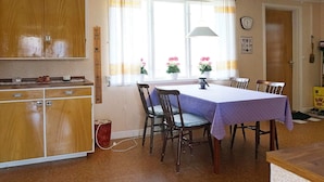 Uma sala de jantar com mesa posta para quatro, armários de madeira, uma janela com cortinas e um relógio na parede.