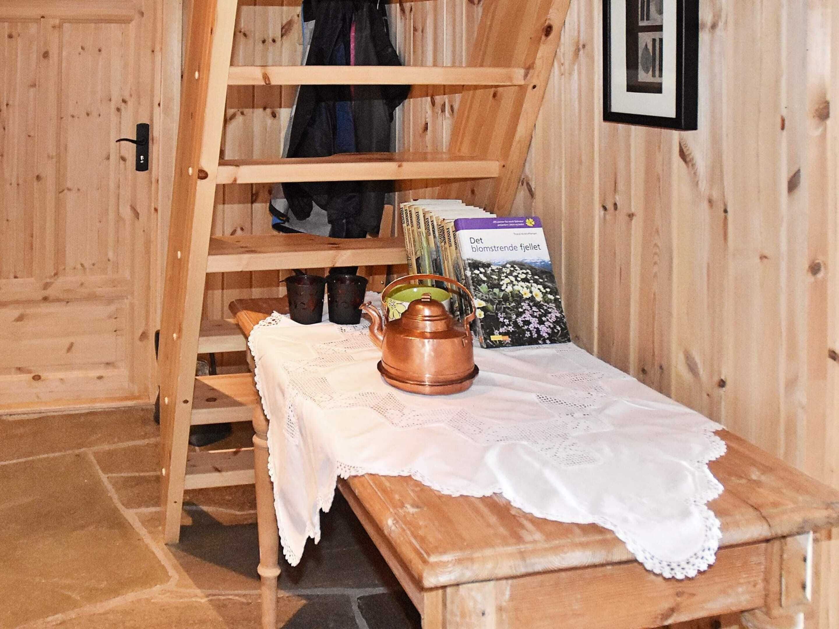 A wooden interior with a bench, a table with a teapot, and a shelf with books and cups.