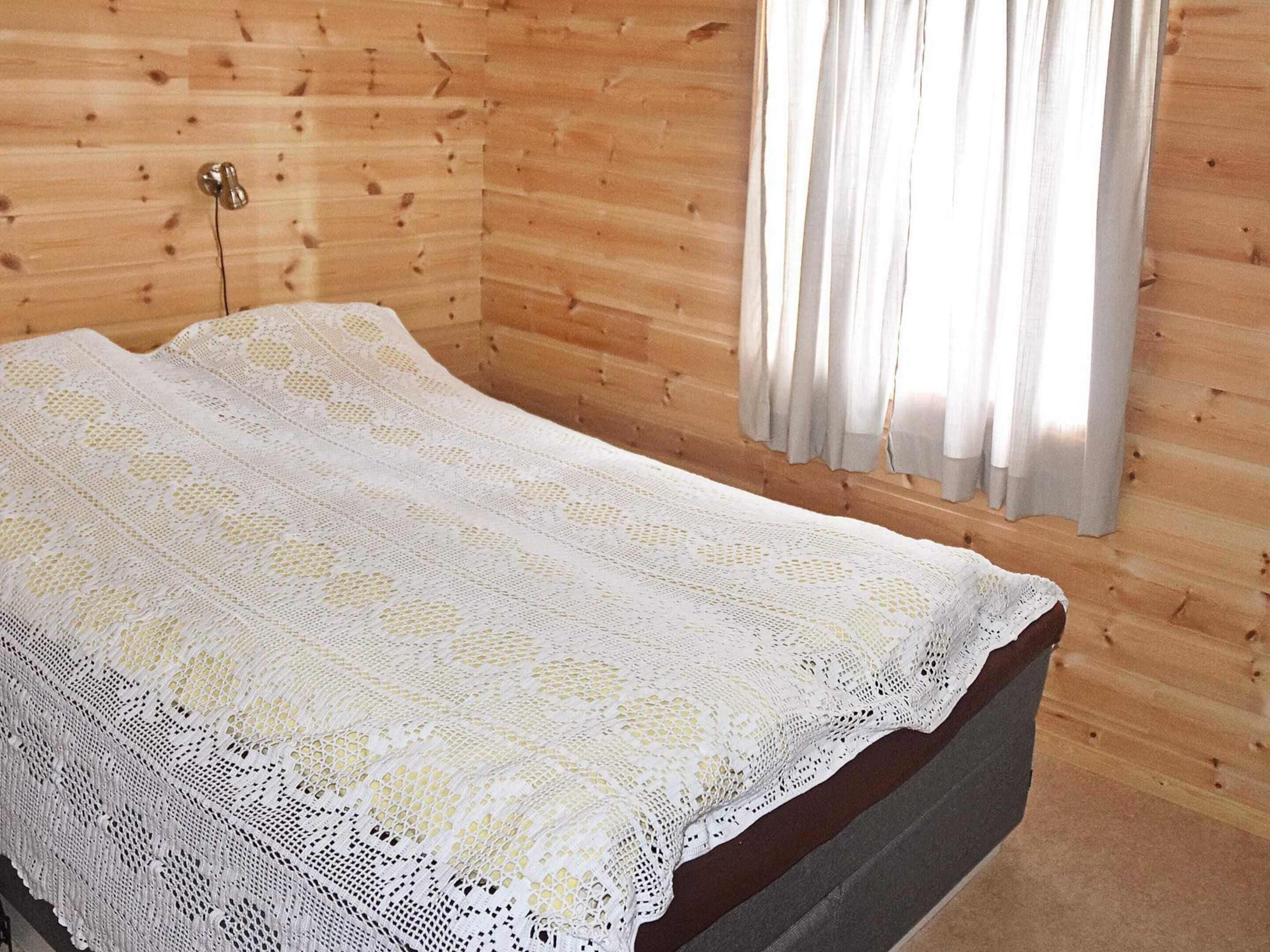 Una cama individual con una colcha de encaje blanco en una habitación con paredes de madera y una ventana con cortinas.