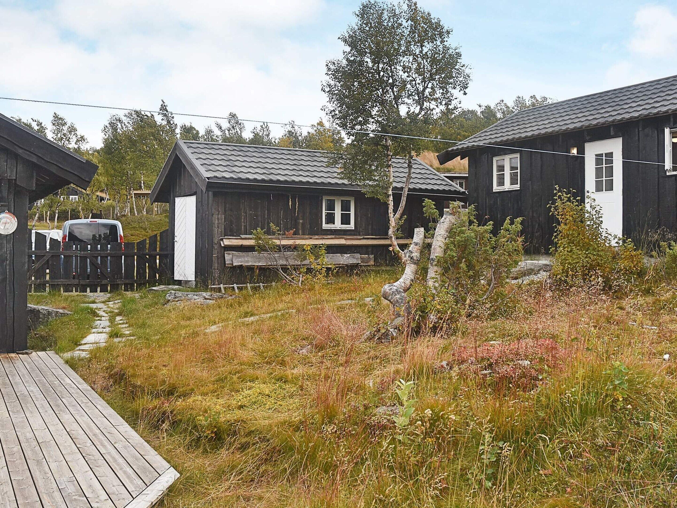 A rustic cabin with a wooden deck, surrounded by natural vegetation and a clear sky.