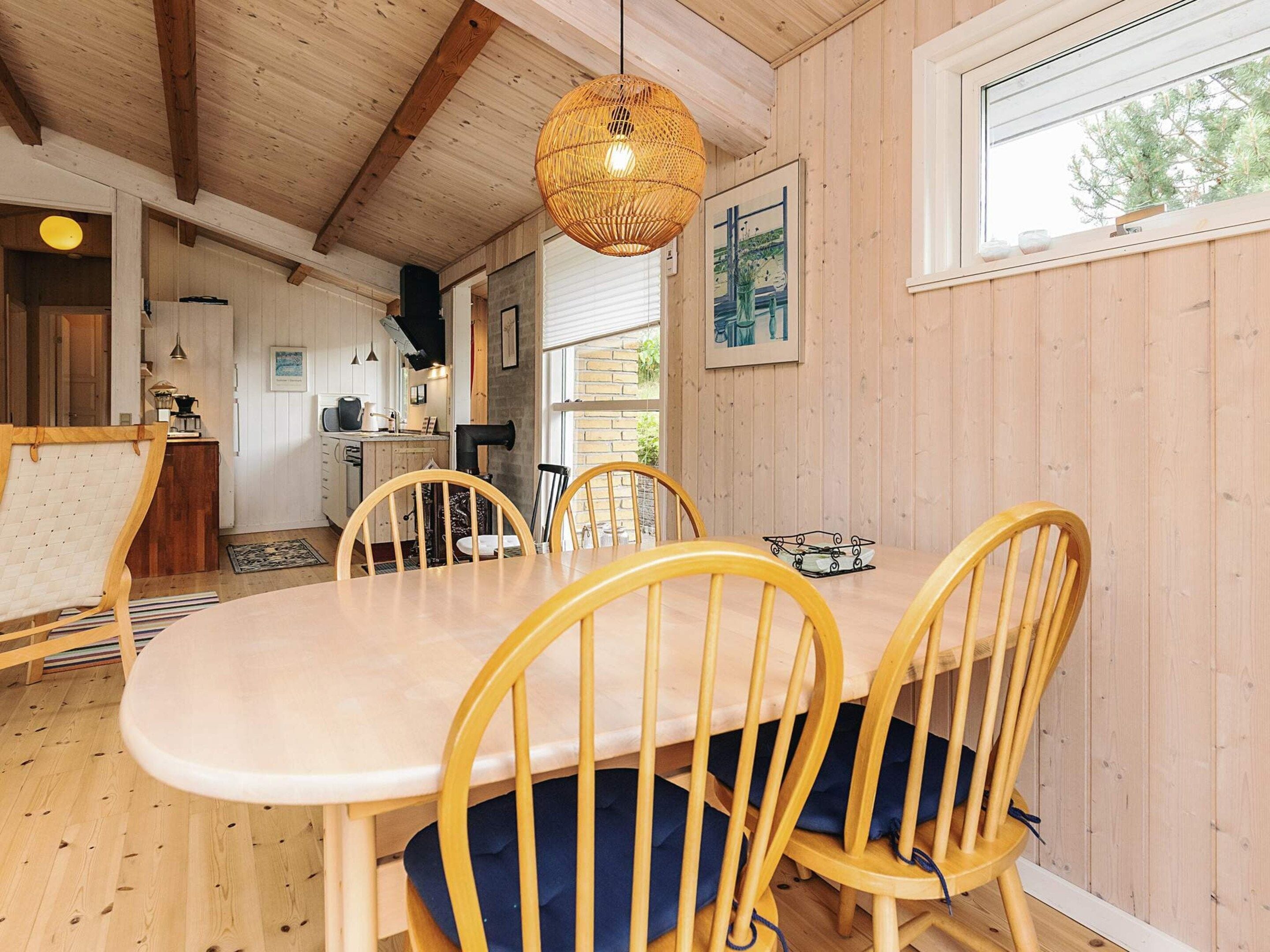Interior de madera con una mesa y sillas de comedor, una lámpara colgante y una ventana que da a vistas de árboles.