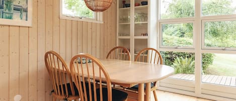 A wooden-paneled room with a round table and chairs, a hanging light fixture, and a view of greenery through large windows.