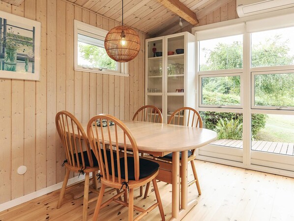 Una habitación con paneles de madera, una mesa redonda y sillas, una lámpara colgante y vistas a follaje a través de amplios ventanales.