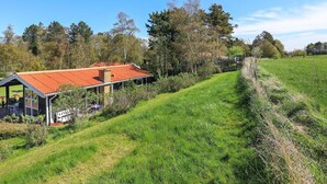 Une maison moderne avec un toit rouge, entourée d’un champ verdoyant et d’arbres.