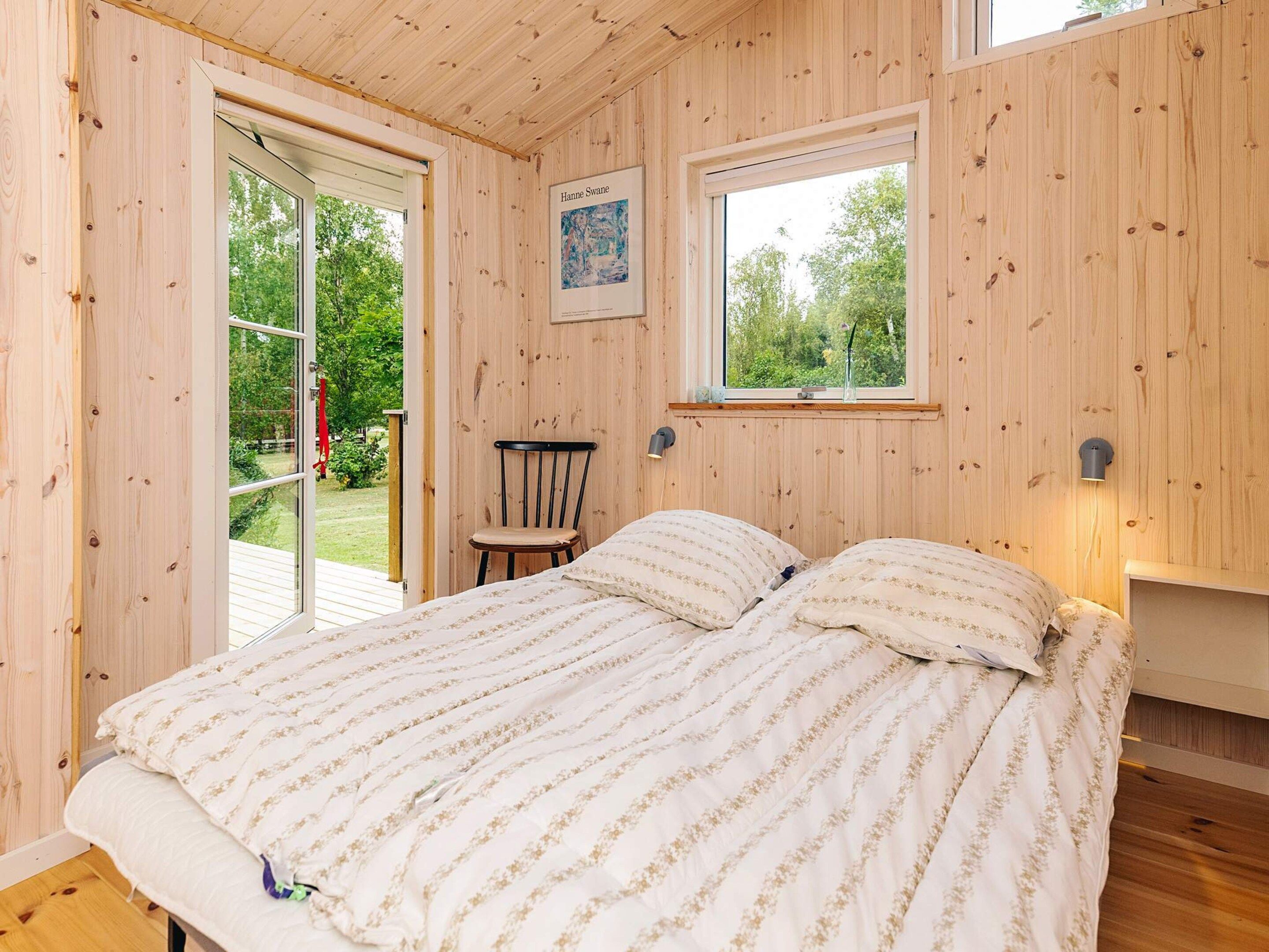 A wooden-paneled bedroom with a bed, two windows, a chair, and a framed picture on the wall.