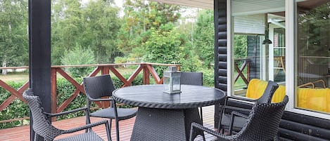 En overdækket terrasse med et rundt bord og fire stole, omgivet af en træterrasse og gelænder af glas.