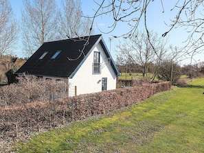 A white house with a black roof, surrounded by a hedge and trees.