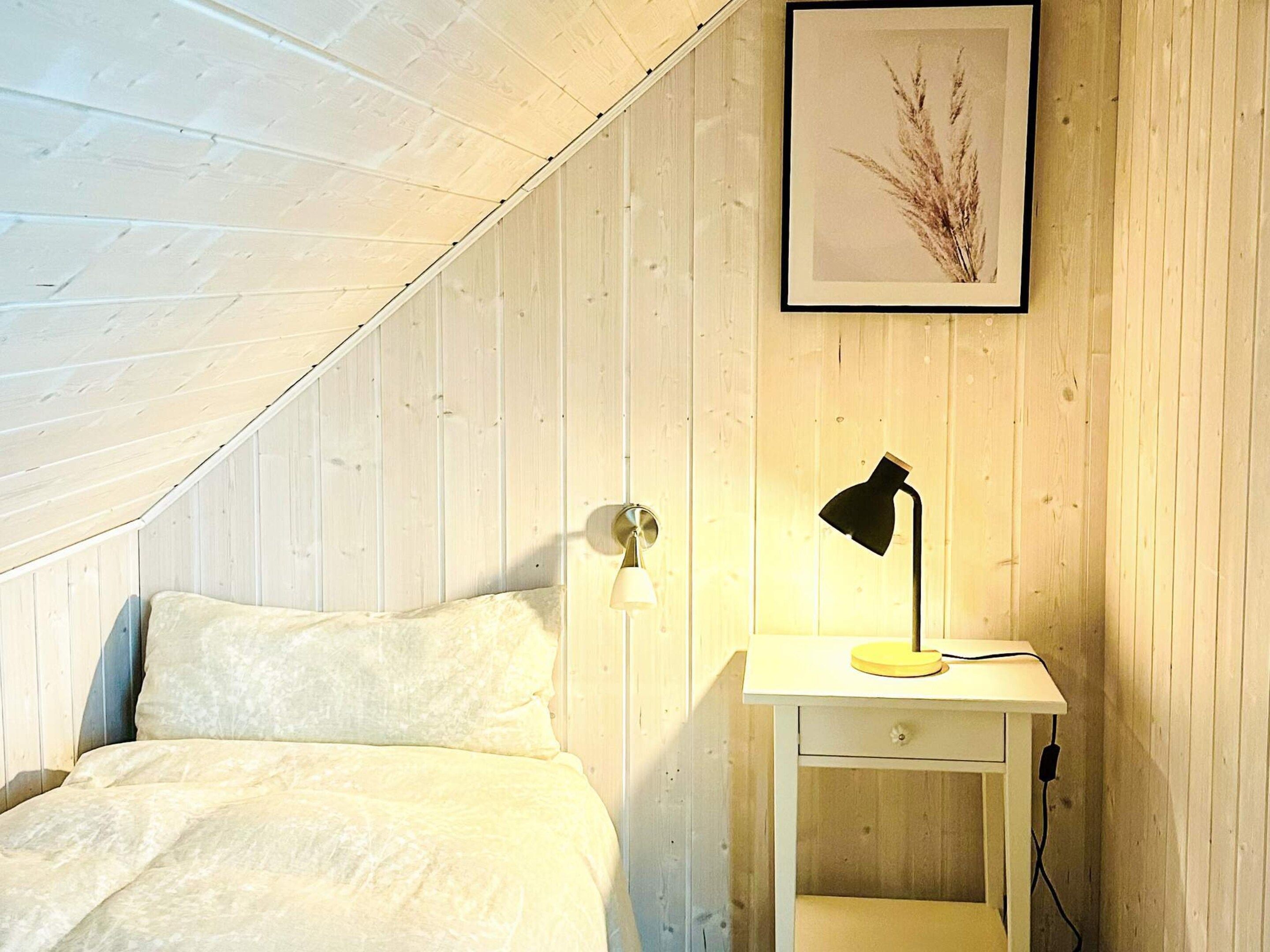 A bedroom with a sloped ceiling, a bed with white bedding, a bedside table with a lamp, and a framed picture on the wall.
