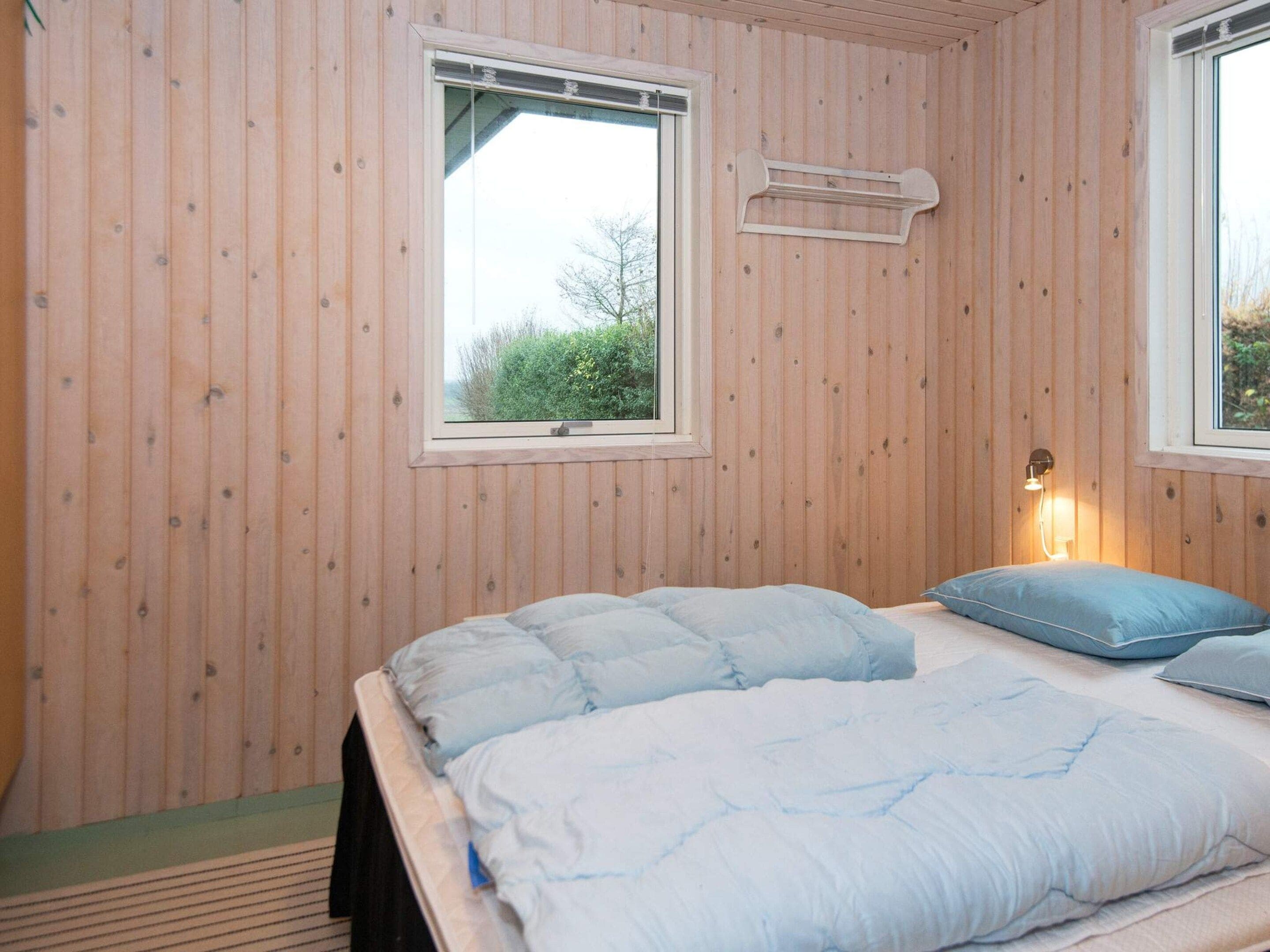 A room with wooden walls, a bed with blue bedding, a window, and a wall-mounted heater.