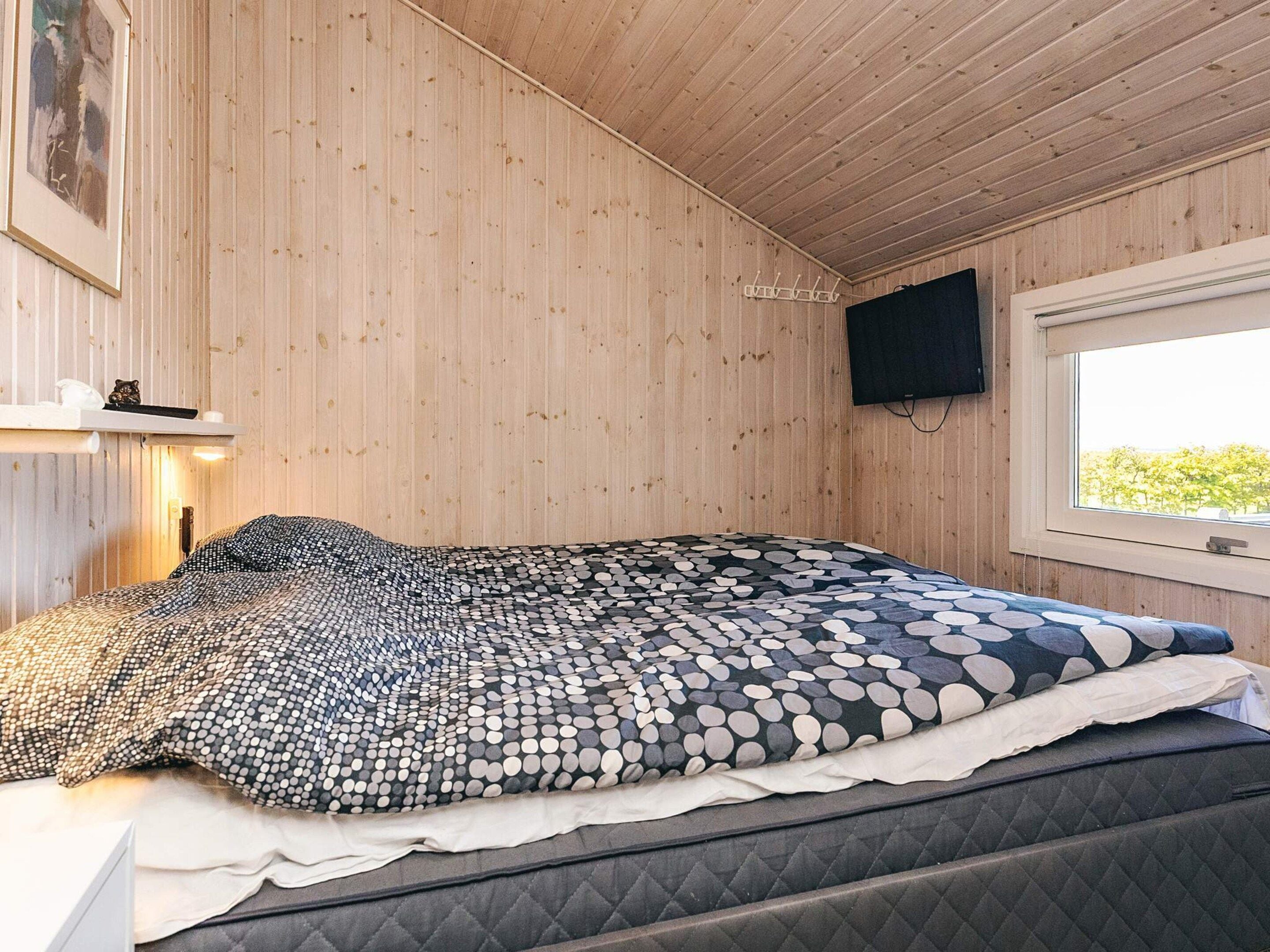 A room with wooden walls, a bed with patterned bedding, a mounted TV, and a window with a view.