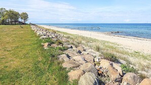 Une plage bordée d’une côte rocheuse, avec une jetée en bois et une zone herbeuse parsemée d’arbres.