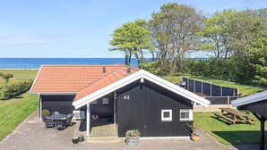 Une maison située sur la côte, avec un toit recouvert de tuiles, le numéro « 44 » peint sur le côté, une terrasse avec des chaises et une table, et une vue sur la mer.