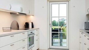 Et moderne køkken med hvide skabe, en indbygget ovn og et stort vindue med udsigt til en have.