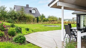 En træterrasse med et bord og stole, omgivet af en velplejet have og et hus i baggrunden.