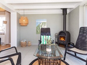 A modern living room with a glass-top table, wicker chairs, a black sofa, a woven hanging light, and a wood-burning stove.