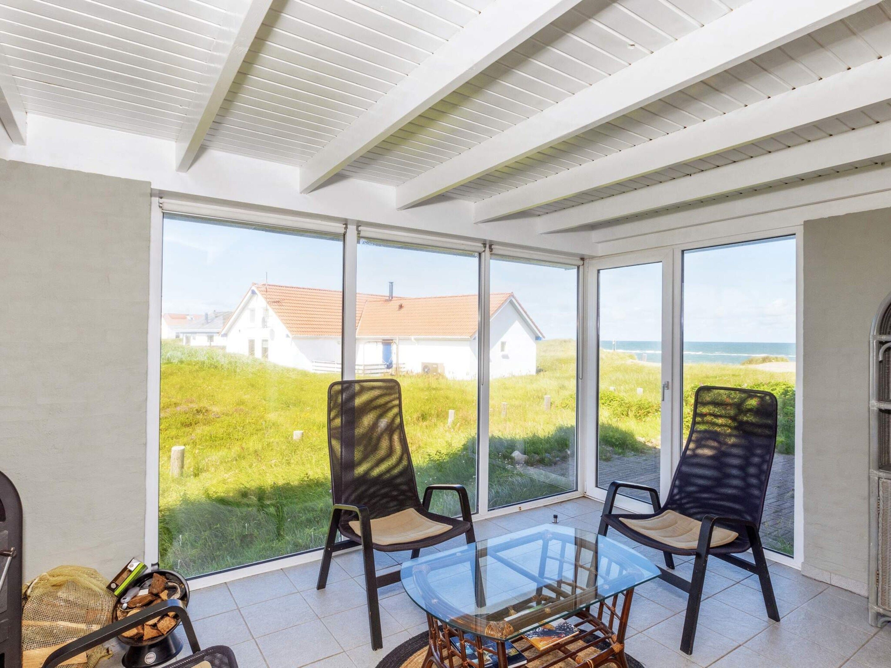 A sunlit room with a glass table and chairs, large windows offering a view of a house and beach.