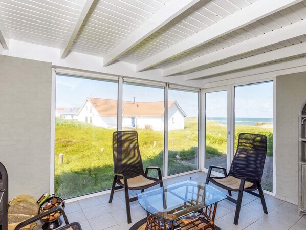 A sunlit room with a glass table and chairs, large windows offering a view of a house and beach.