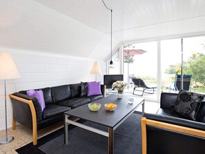 A modern living room with a black leather sofa, a glass coffee table, and a dining table. There is a large window providing a view of the outdoors. - Quaint Holiday Home in Sjaellands Odde near Sea (Sjællands Odde)