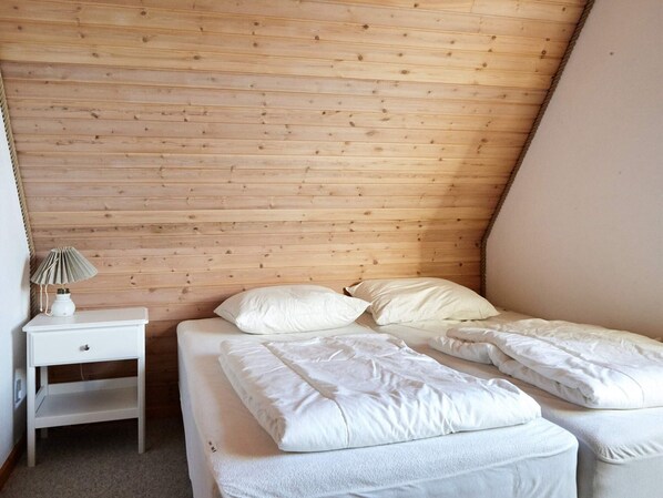 Una habitación pequeña y acogedora con techo inclinado y paneles de madera. Cuenta con una cama individual con ropa de cama blanca, una mesita de noche con lámpara y un estante pequeño.