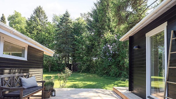 A wooden deck with a bench and potted plants, surrounded by a lush garden and a dark wooden building with a glass door. - 6 Person Holiday Home in Gilleleje-by Traum (Gilleleje)