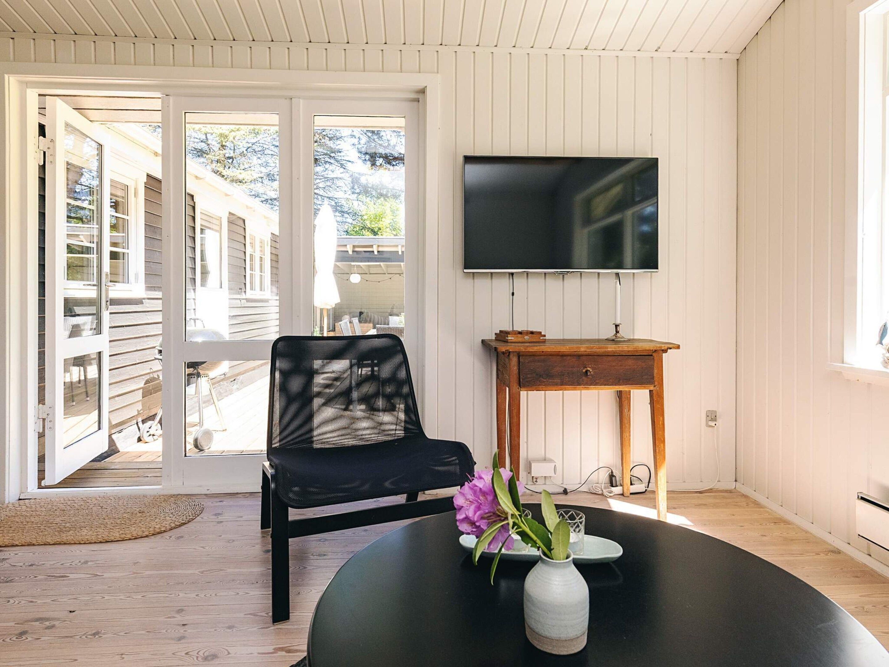 A room with a black chair, a round table with a vase of flowers, a wooden side table, and a TV mounted on the wall.