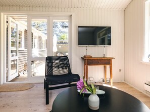 Une pièce comprenant une chaise noire, une table ronde ornée d’un vase de fleurs, une table d’appoint en bois et un téléviseur fixé au mur.