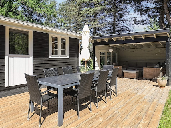 A wooden deck with a dining table and chairs, a covered seating area, and a small house with a white door and windows.