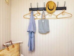 Une pièce avec un mur blanc, un porte-manteau noir sur lequel sont accrochés des chapeaux et des vêtements rayés, un panier en osier et une chaise blanche.