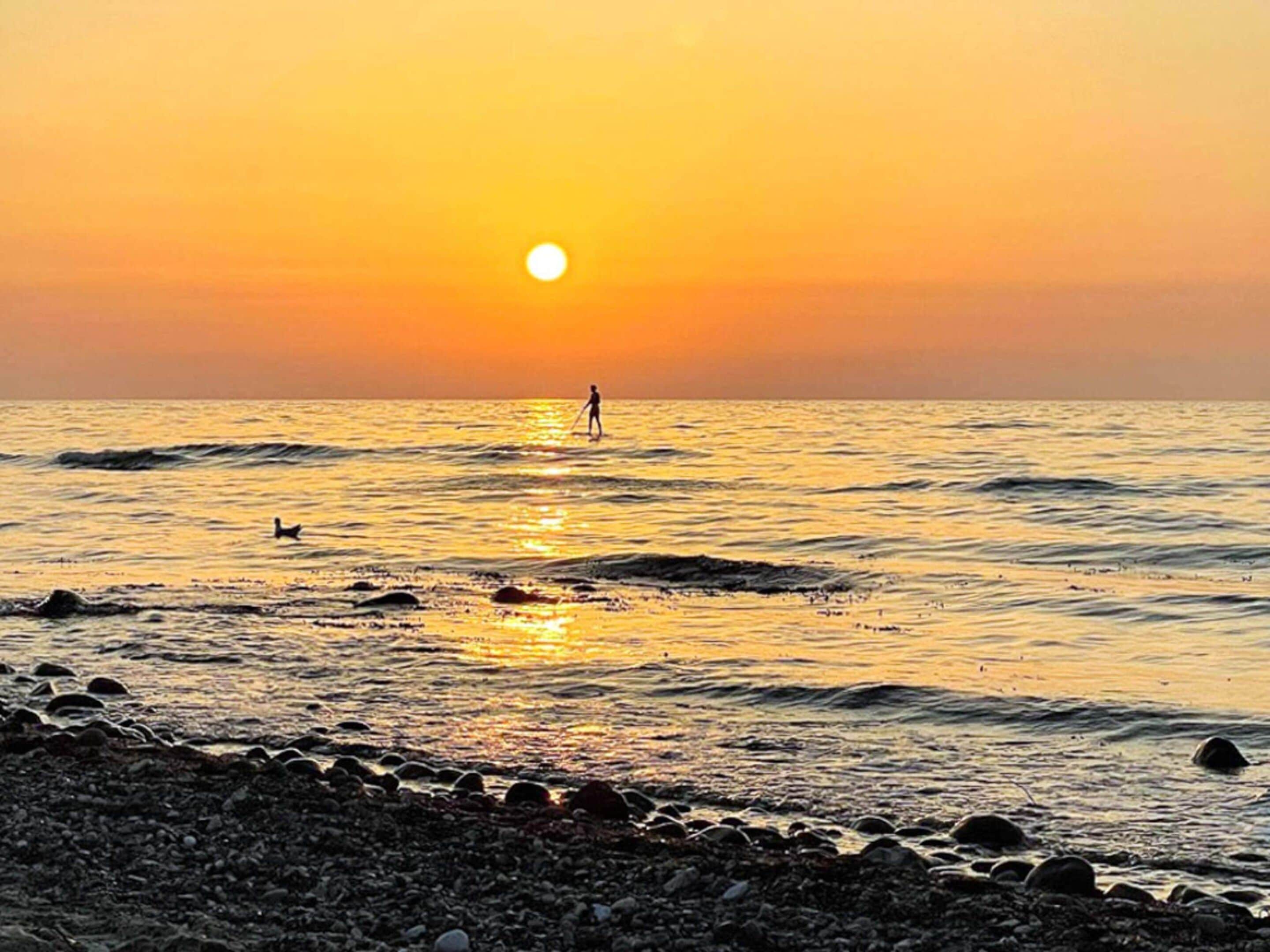 A sunset over a rocky beach with waves and a person standing in the water.