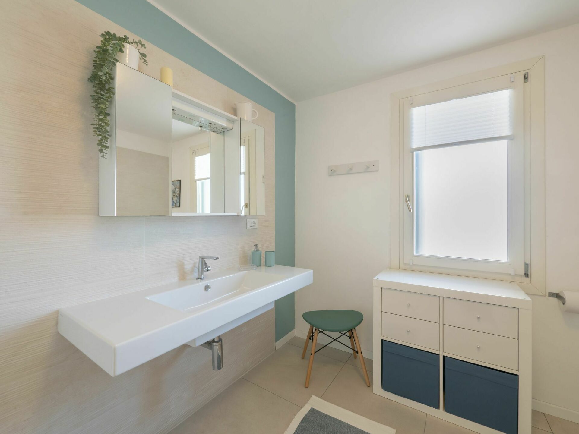 Ein Badezimmer mit Waschbecken, Spiegel, Fenster und einem kleinen Hocker.