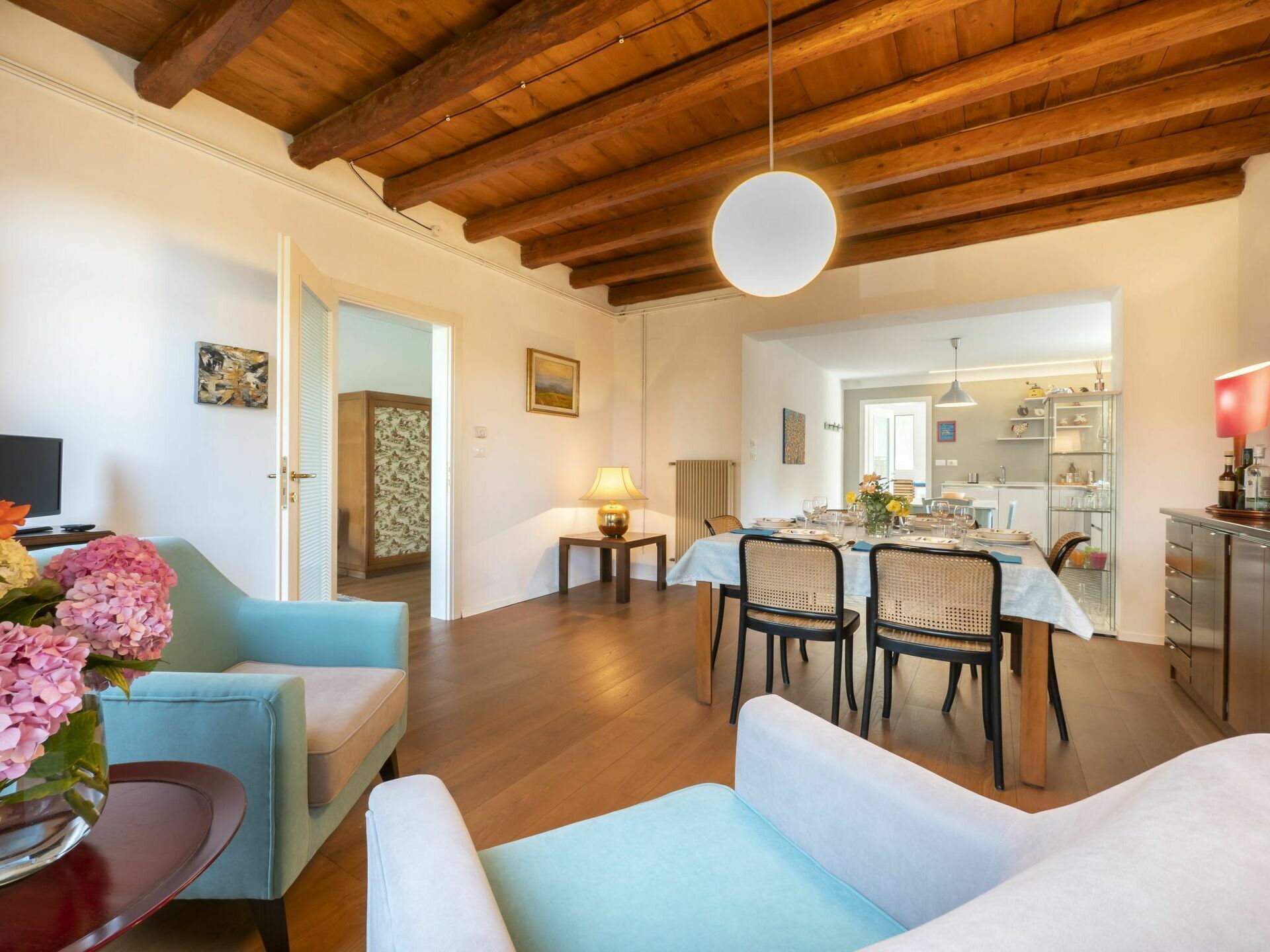 A spacious living room with a wooden ceiling, a dining area with a table set for four, and a kitchen visible in the background.