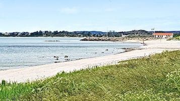 En strand med en gräsklädd förgrund, en sandstrand och en byggnad med ett rött tak i bakgrunden.