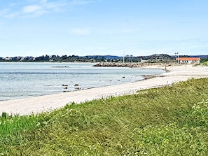 A coastal landscape with a sandy beach, a grassy foreground, and a distant shoreline with buildings. - Holiday Home in Stråvalla (Stråvalla)