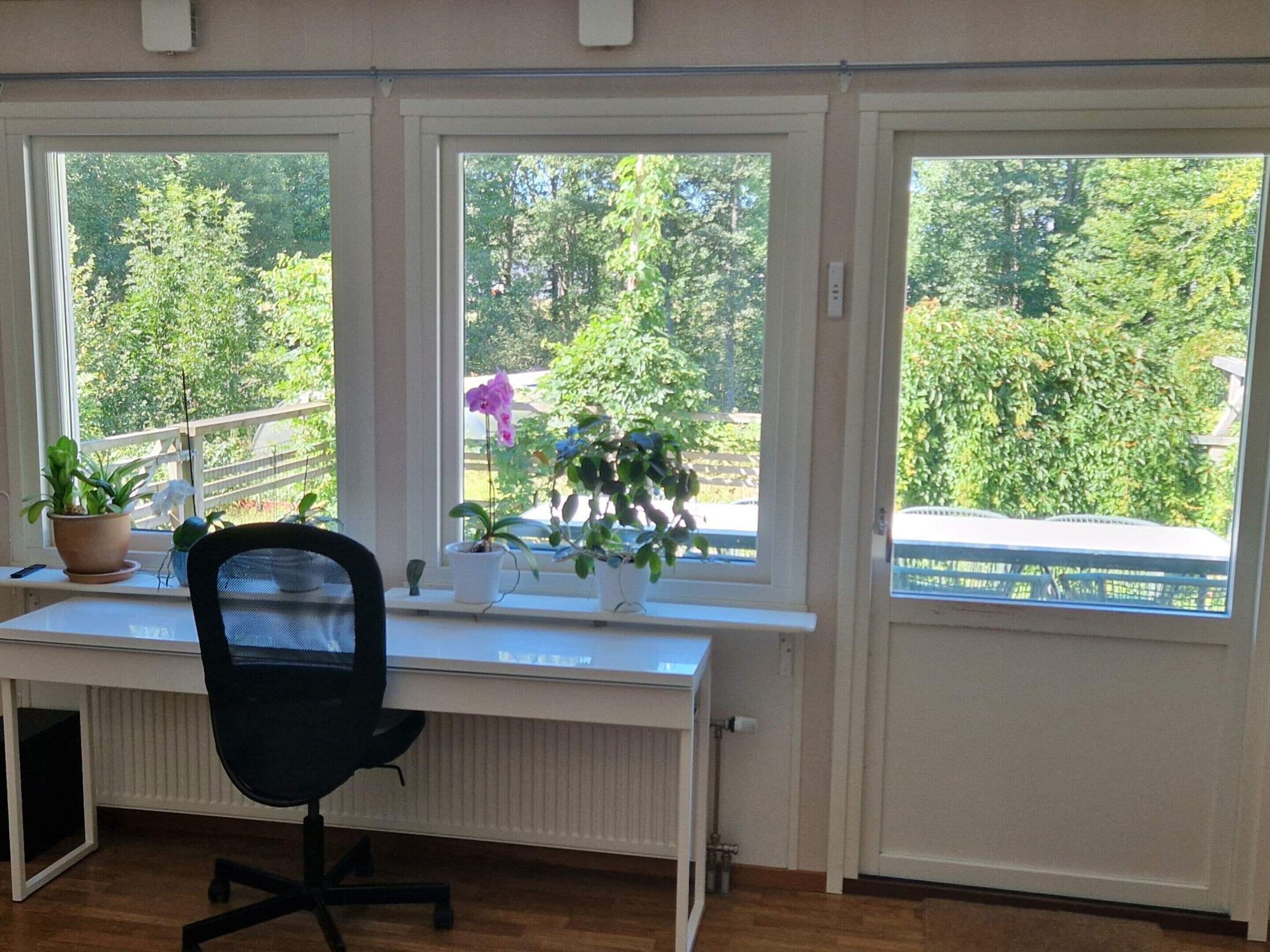 A room with a white desk, black office chair, and three windows overlooking greenery.