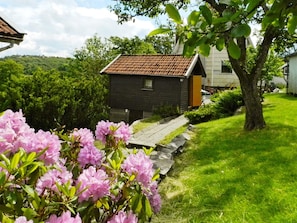 Ett litet hus med ett rött tegeltak, omgivet av en välskött trädgård med rosa blommor och en gång av sten.