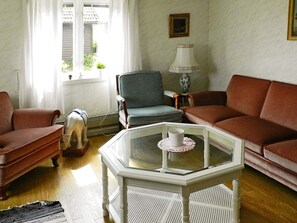 Un salon avec un canapé marron, un fauteuil bleu, une table basse en verre surmontée d’un chemin de table blanc et une petite statuette de chien.