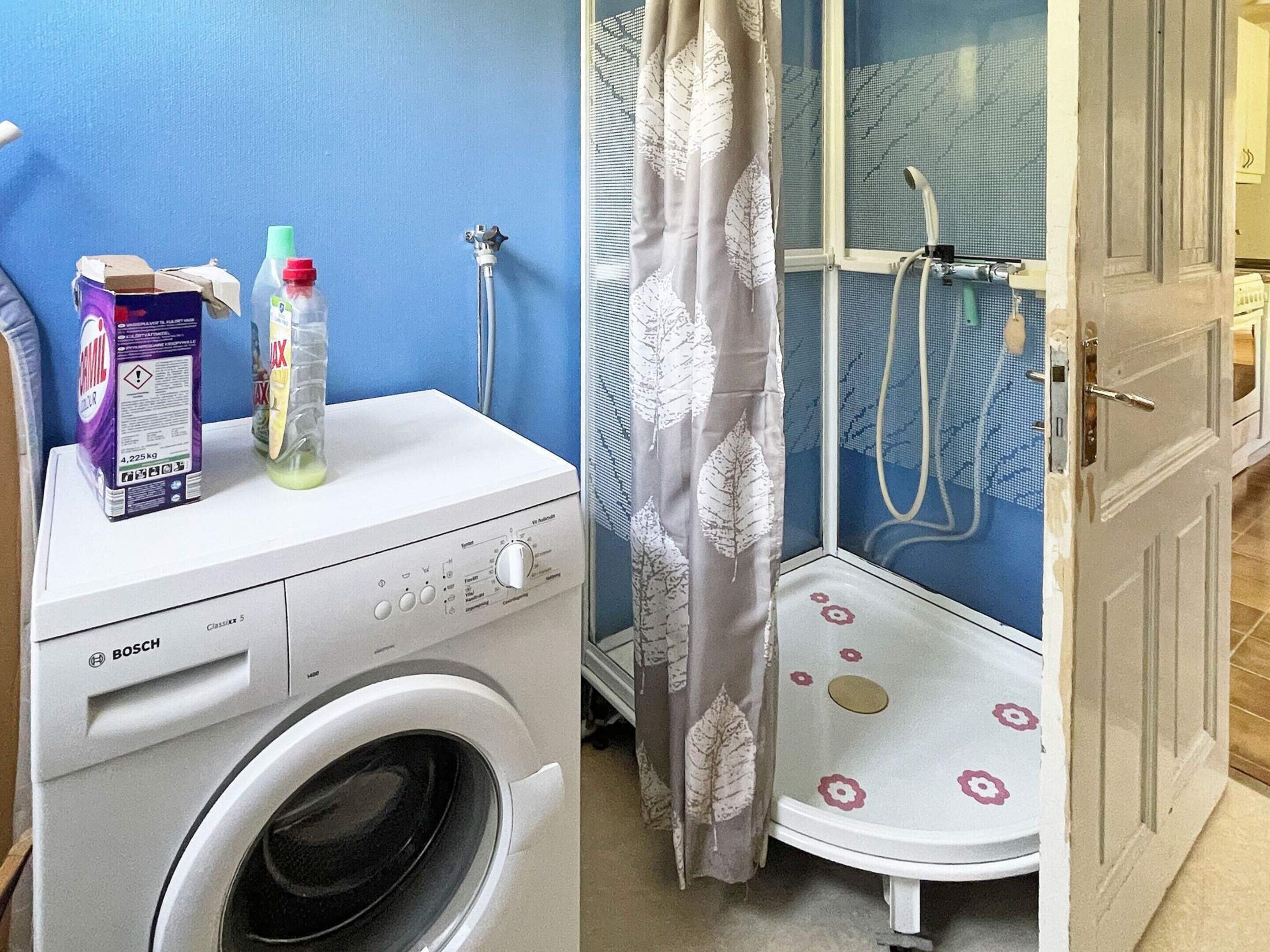 A compact laundry area with a Bosch washing machine, a shower with a floral shower curtain, and a door leading to another room.