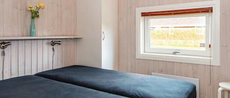 A room with two beds, a window with a view of a field, and a shelf with flowers.