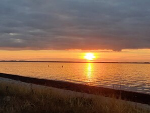 Solnedgang over vand med en fjern kystlinje og overskyet himmel.