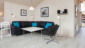 A modern living room with a black sectional sofa, a white coffee table, and a black bar stool. There are two wall-mounted light fixtures and a ceiling light. - Holiday Home in Großenbrode (Grossenbrode)