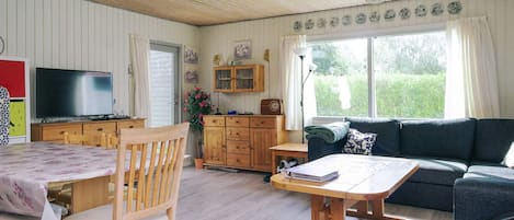 A cozy living room with a wooden ceiling, a television, a dining table with chairs, a sofa, and a window with curtains.