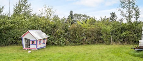 A small, colorful house with a pitched roof and a door, situated in a grassy field with dense bushes and trees in the background.
