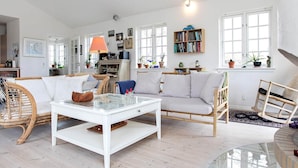 Un salón espacioso con un sofá blanco, una mesa de centro de vidrio y un sillón de mimbre. Hay una estantería llena de libros y una lámpara sobre una mesita auxiliar.