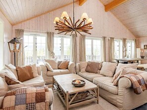 A cozy living room with a high ceiling, wooden beams, and a central chandelier. There are two sofas and a coffee table with a candle.