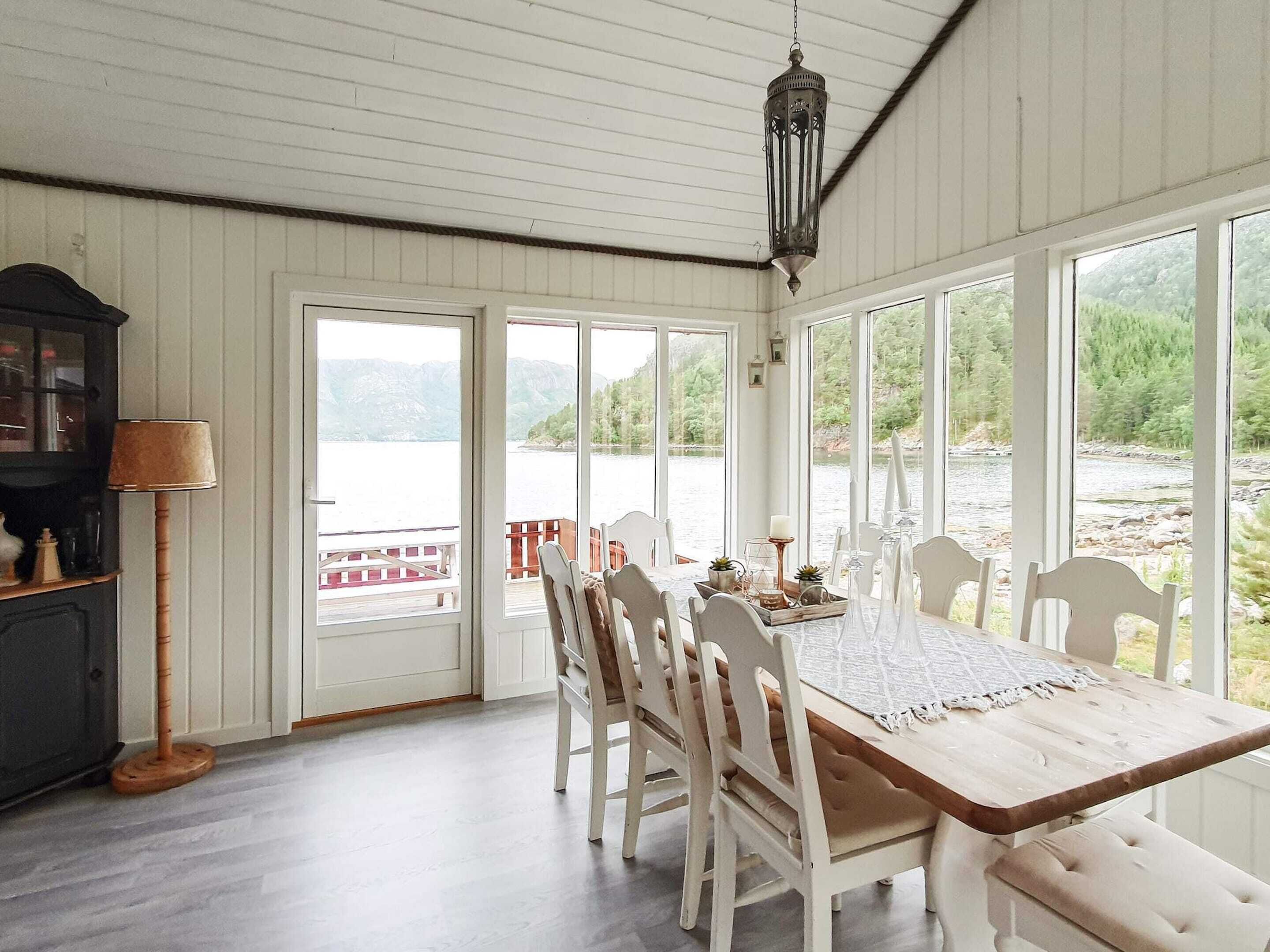 Un comedor con una mesa de madera y sillas blancas, ventanas amplias con vistas a un lago y montañas, y una lámpara colgante.