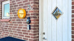 En mur av tegelstenar med en brevlåda, en dörr med en rombformad spegel och ett fönster med utsikt över träd.