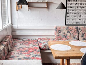 Ein Zimmer mit einer hölzernen Bank mit floralem Bezug, einem Holztisch mit Marmorplatten und einem an der Wand montierten Regal mit diversen Gegenständen.
