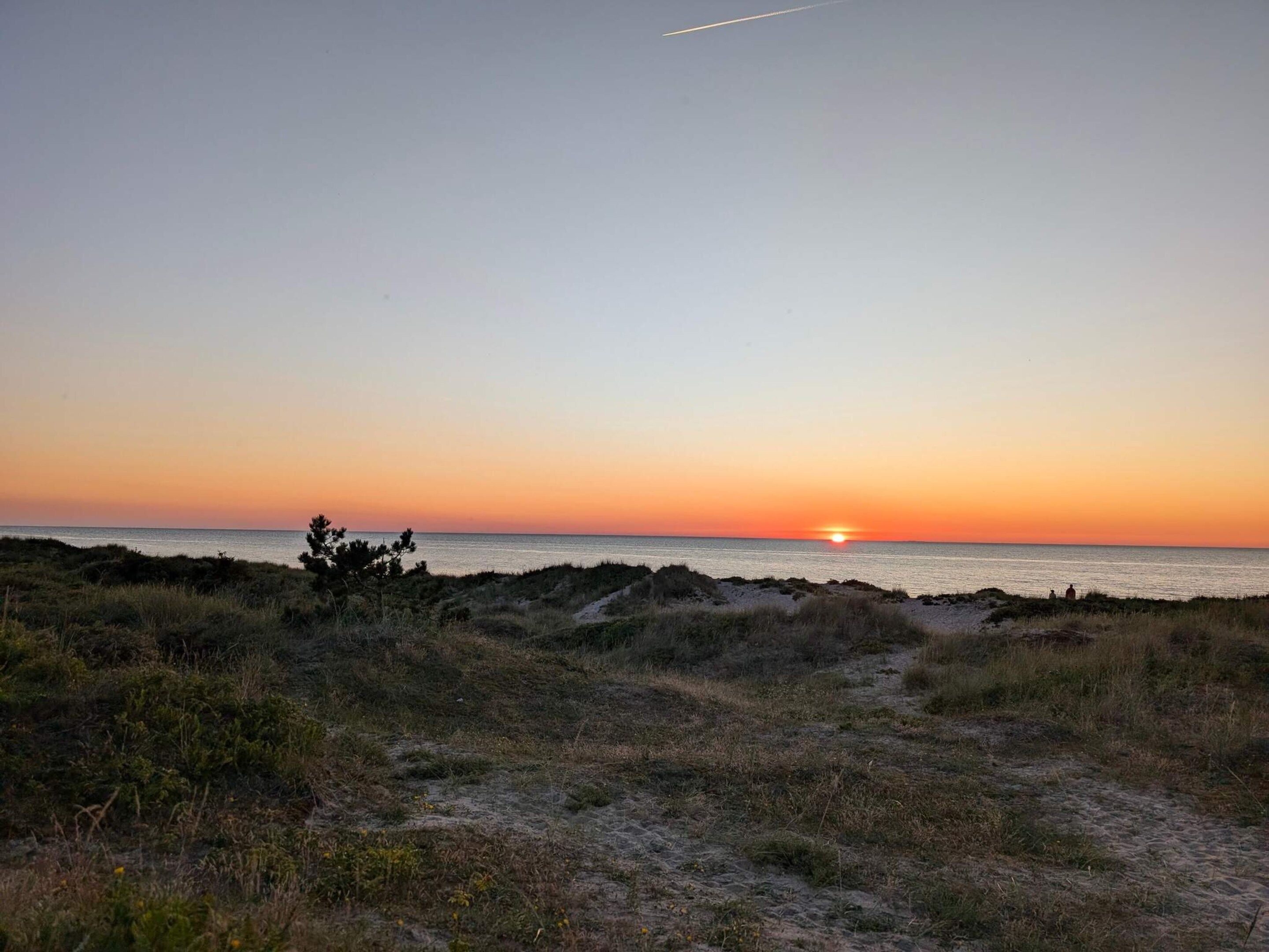 A coastal landscape with a sunset over the ocean, sandy terrain, and sparse vegetation.